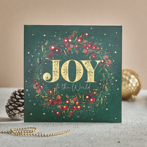 A Christmas card with a Christmas wreath against a green background. The text on the card reads 'Joy to the World'.