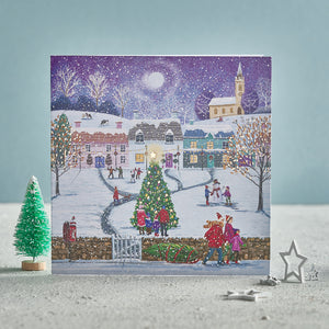A Christmas card showing a tree in a village with people all around wrapped up in the snow