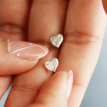 Load image into Gallery viewer, Heart-shaped silver earrings held between fingers against a neutral background